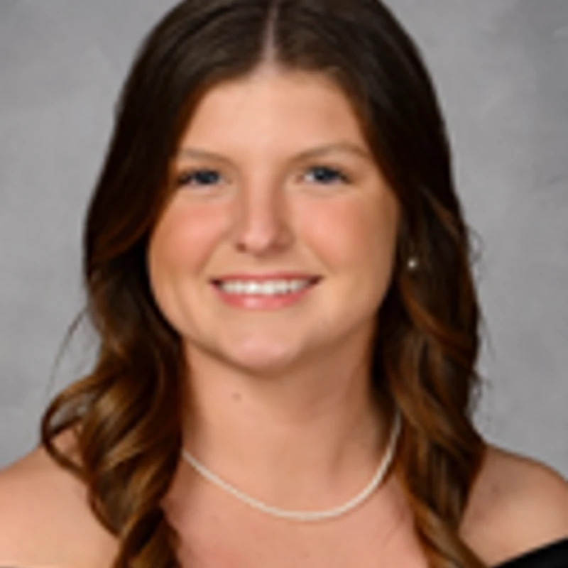 A formal headshot of a young woman with long, wavy brown hair. She is smiling and wearing a black off-the-shoulder top and a pearl necklace.