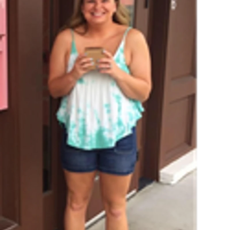 A smiling blonde woman stands outside a storefront, wearing a turquoise tie-dye tank top and denim shorts while holding a small brown cup.
