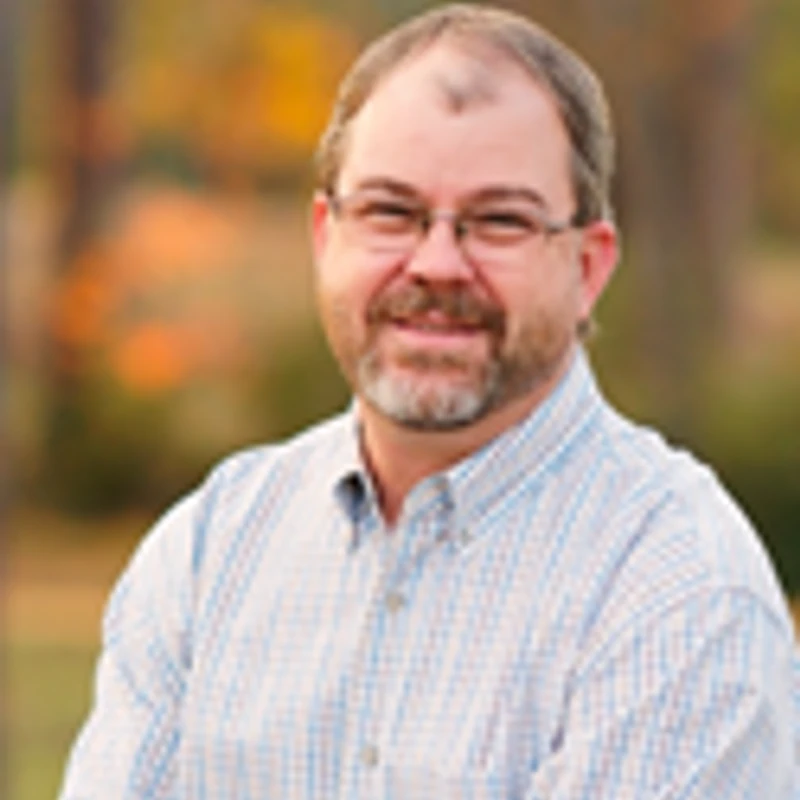 A headshot of a smiling, middle-aged man with glasses and a beard, wearing a plaid button-down shirt. The background is a soft-focus view of autumn trees.