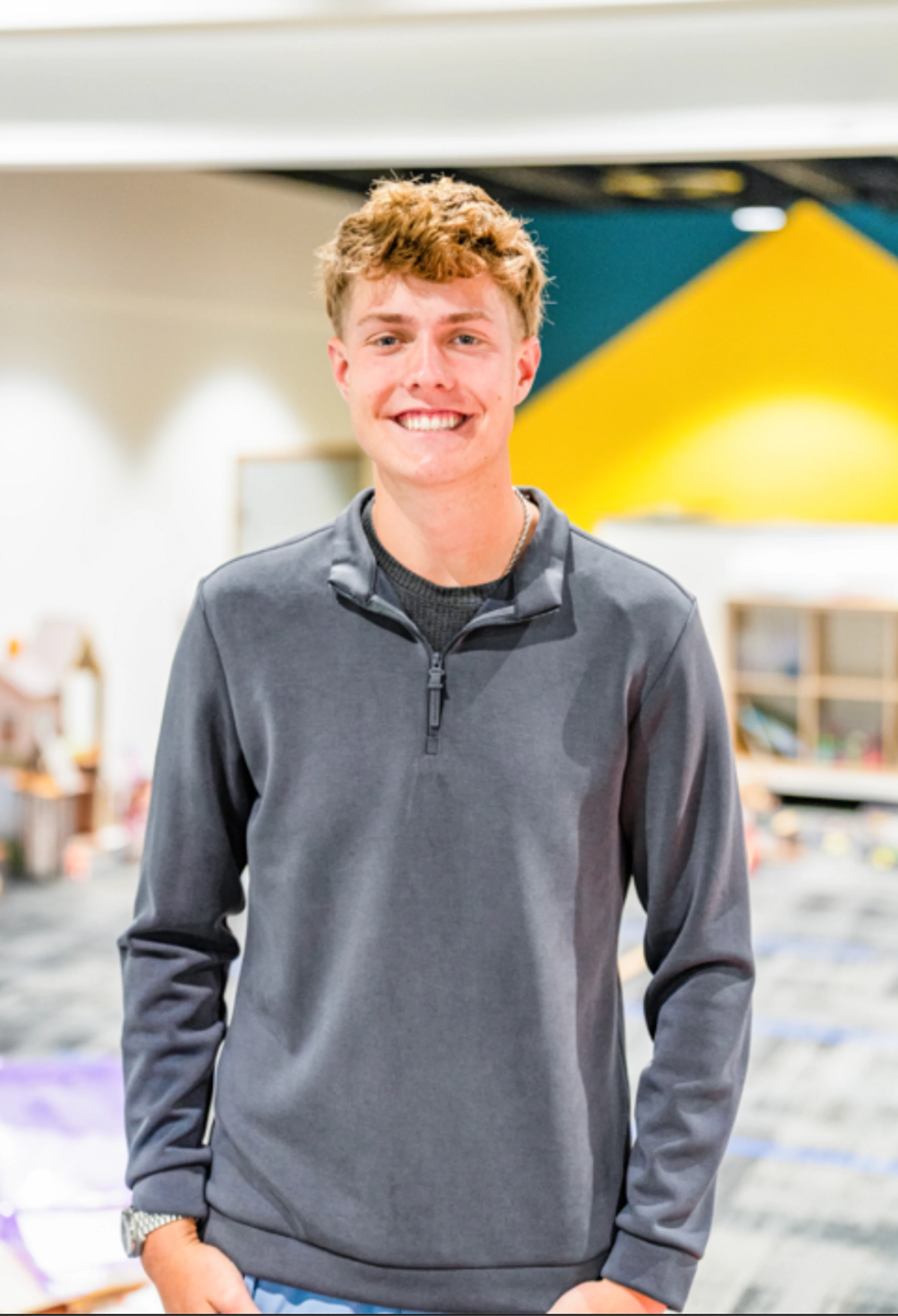 A young man with curly blond hair wearing a grey quarter-zip sweatshirt smiles at the camera with his hands in his pockets while standing indoors.