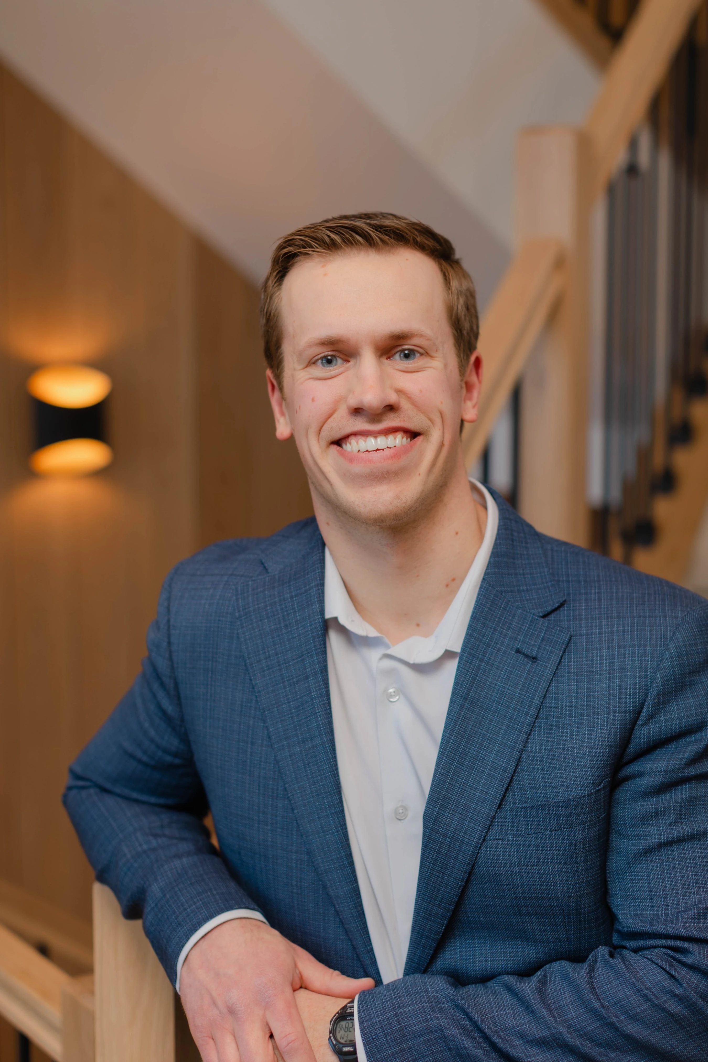 A young man with short brown hair and blue eyes smiles warmly. He is wearing a blue plaid suit jacket over a white shirt and leaning on a wooden railing.