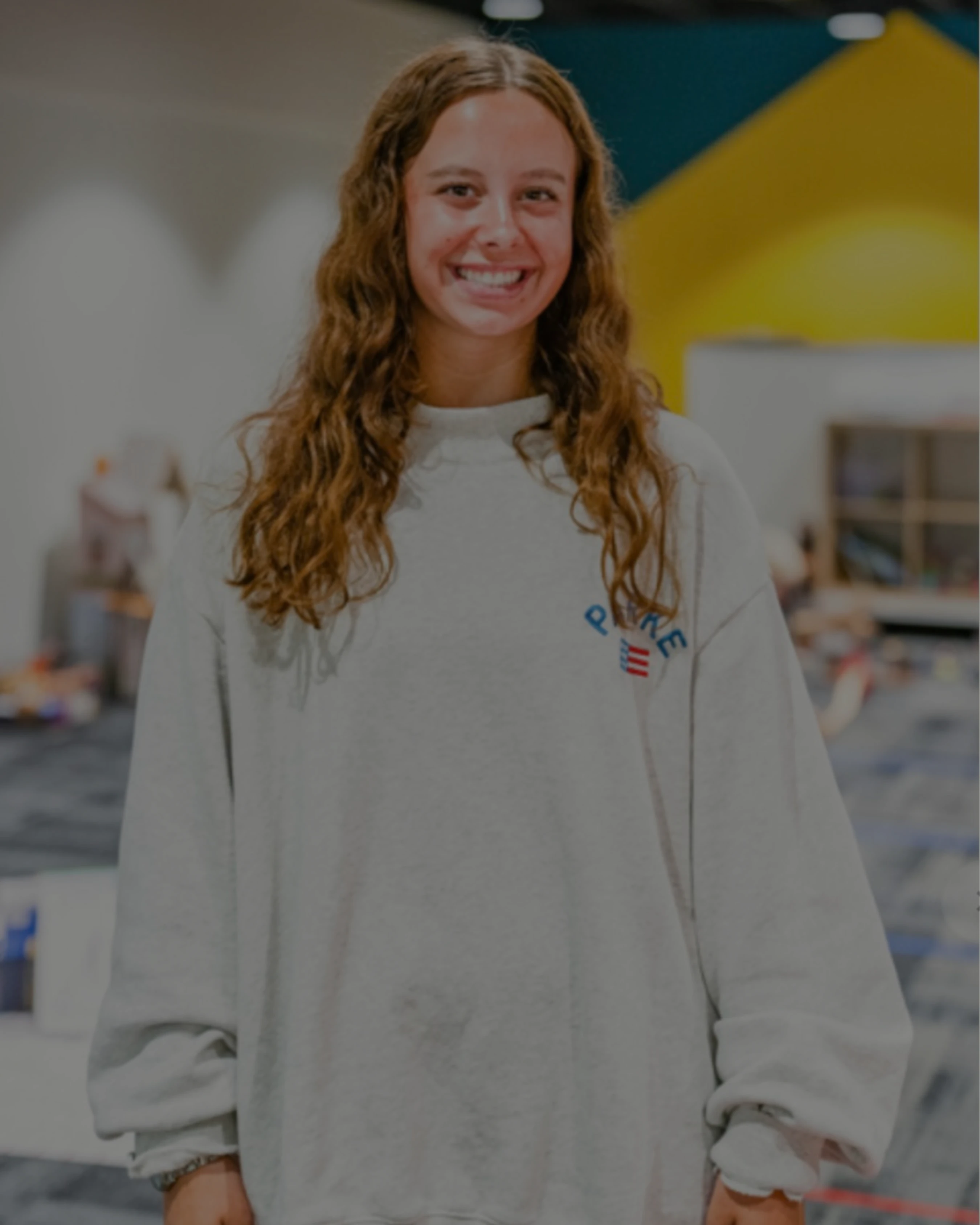 A young woman with long, wavy brown hair stands indoors and smiles at the camera. She is wearing a light gray oversized sweatshirt with a small logo.