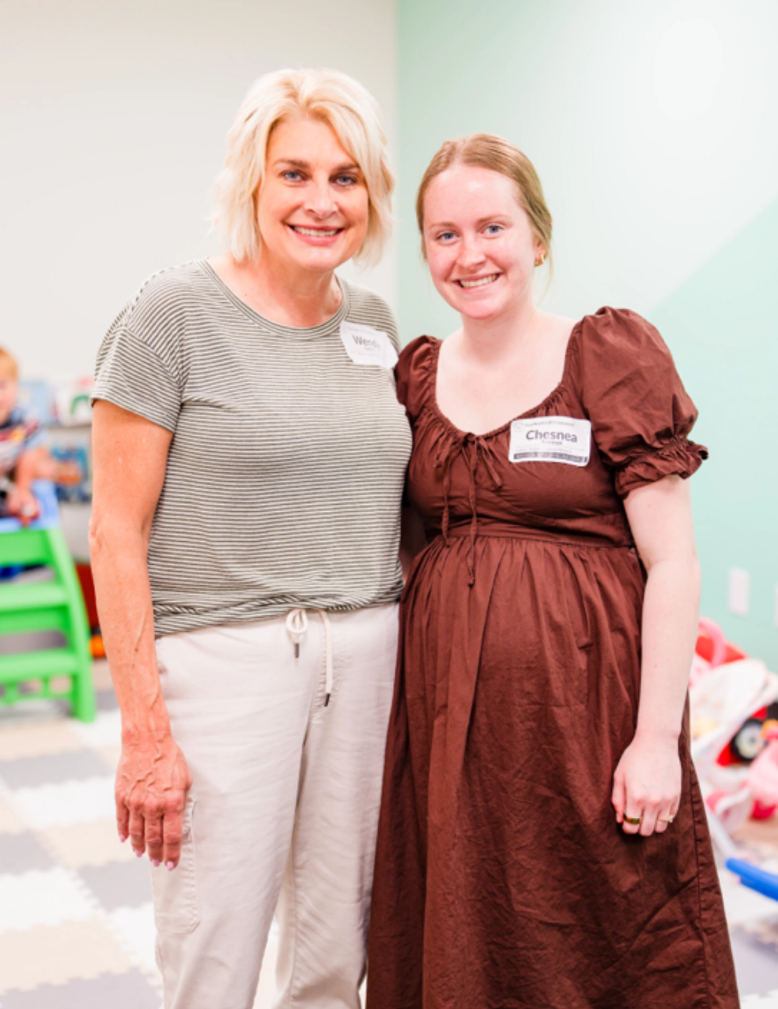 A woman with short blonde hair and a pregnant woman in a brown dress stand side-by-side, smiling for the camera. Both are wearing nametags.
