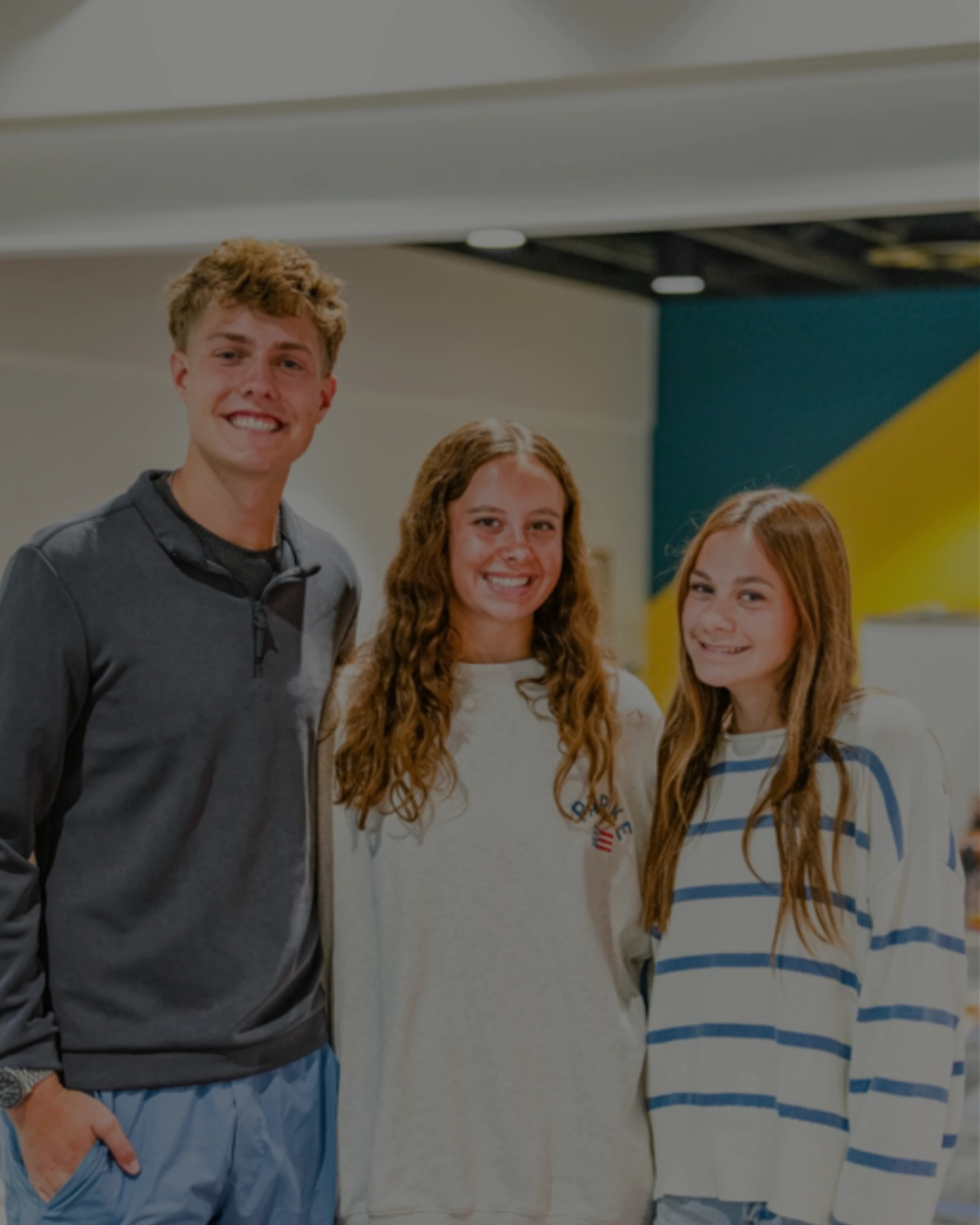 A young man and two young women stand together indoors, smiling happily for the camera. They are all dressed in casual sweaters and sweatshirts.