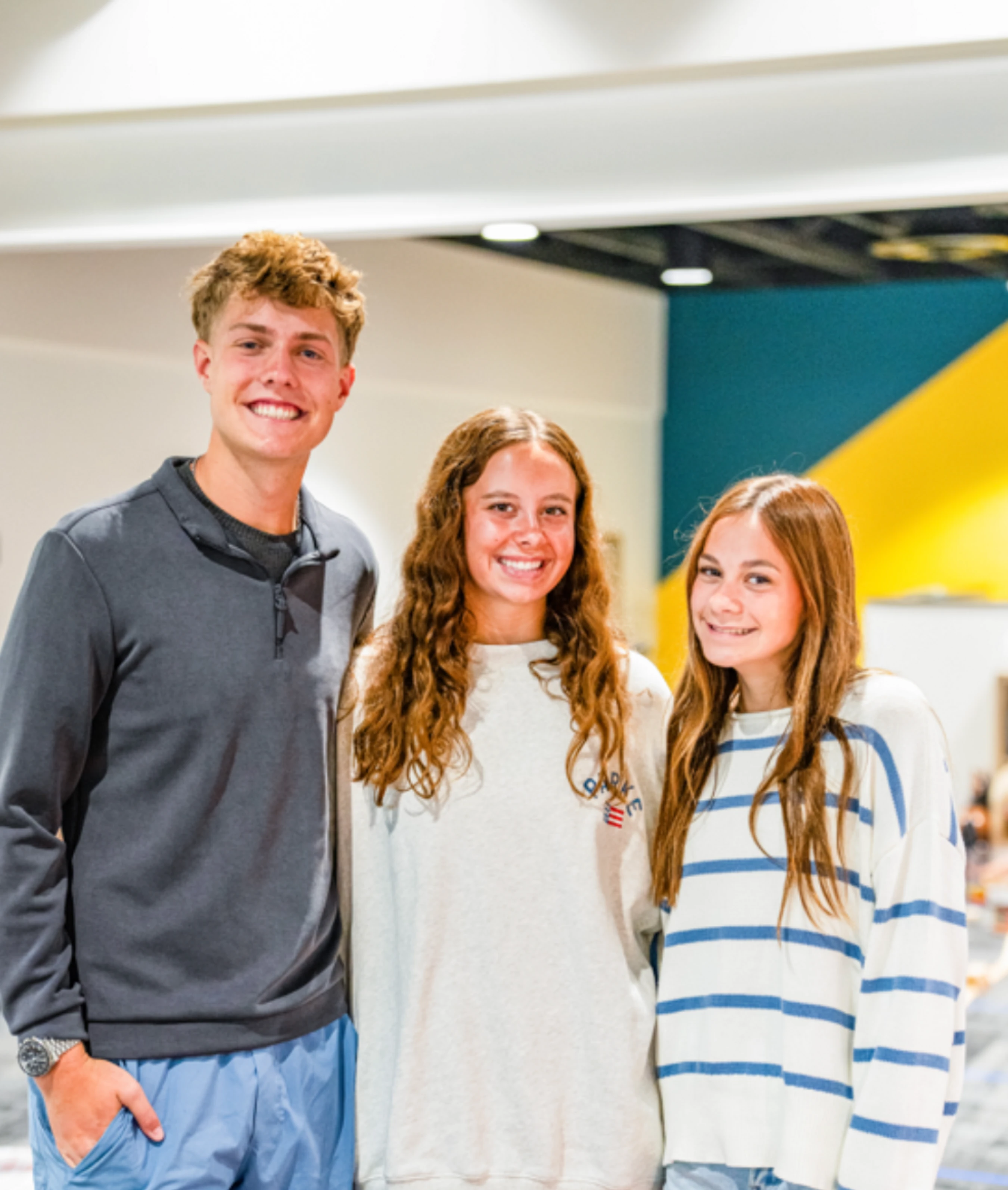 A young man and two young women stand close together, smiling at the camera in a brightly lit room with a white, blue, and yellow background.