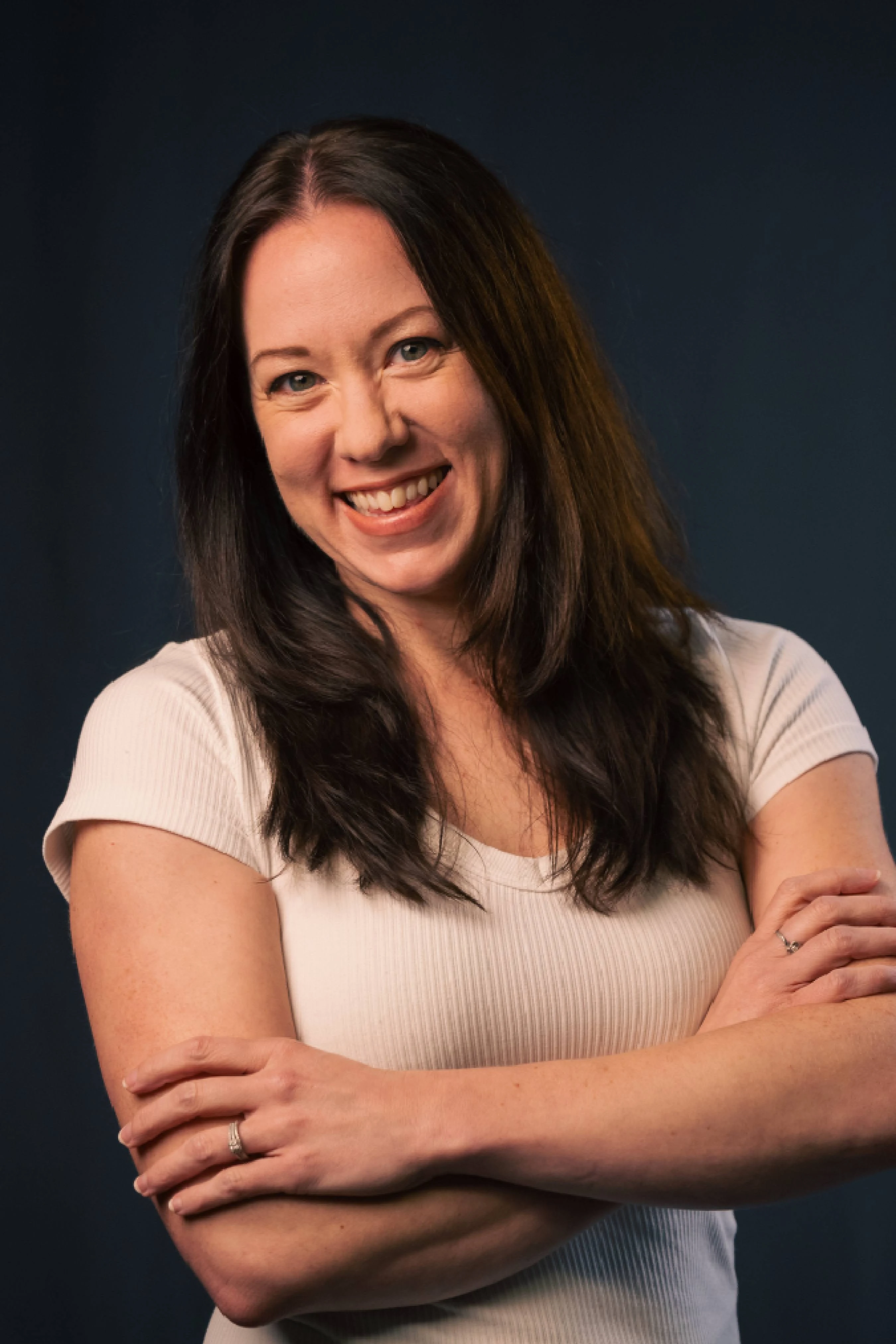 A headshot of a woman with long dark hair, smiling warmly at the camera. She wears a white shirt and has her arms crossed against a dark blue background.