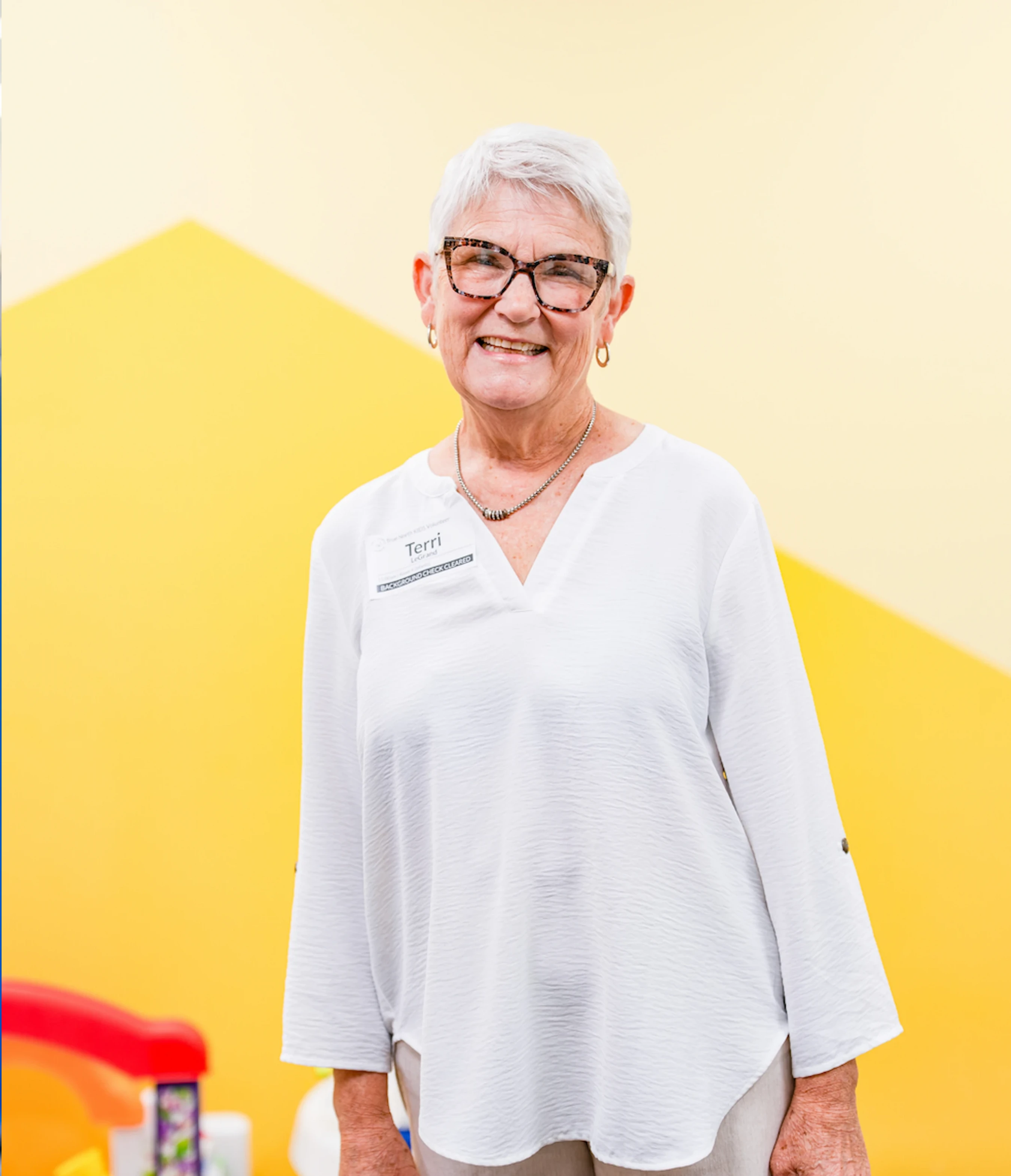A smiling older woman with short white hair and glasses wears a white shirt and a name tag against a two-toned yellow background.