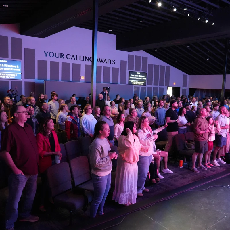 A large, diverse congregation stands and sings in a modern auditorium during a church service, illuminated by purple stage lights. Screens show song lyrics.