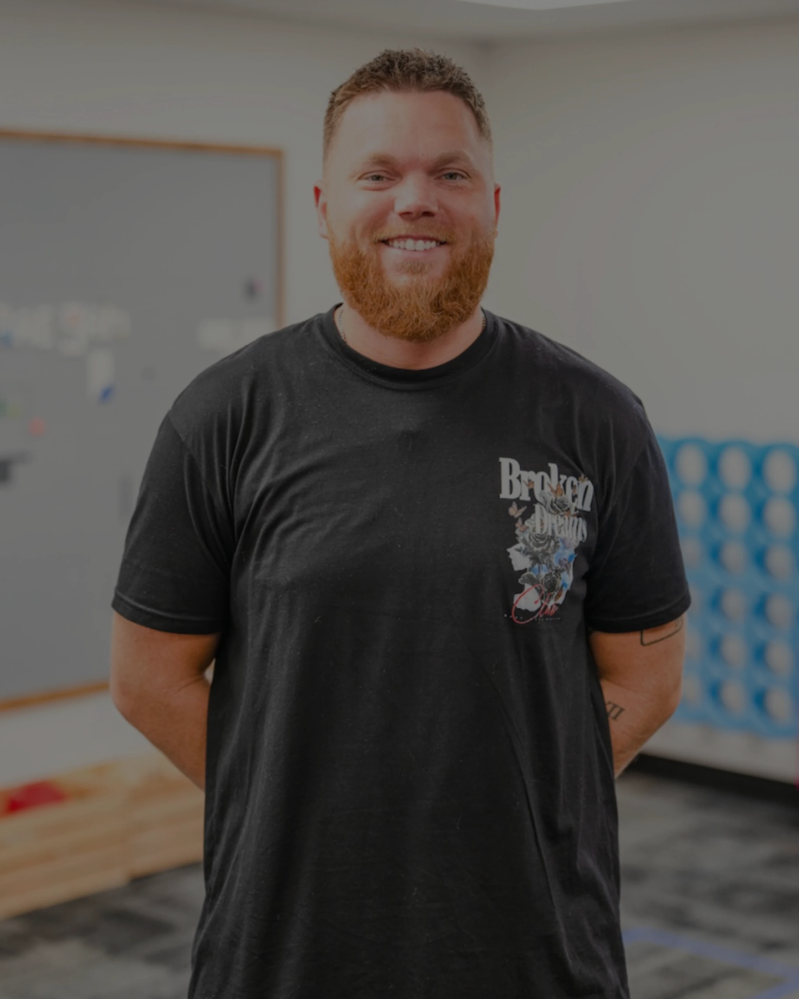 A man with short brown hair and a full reddish beard smiles while standing indoors. He is wearing a black t-shirt and has his hands behind his back.