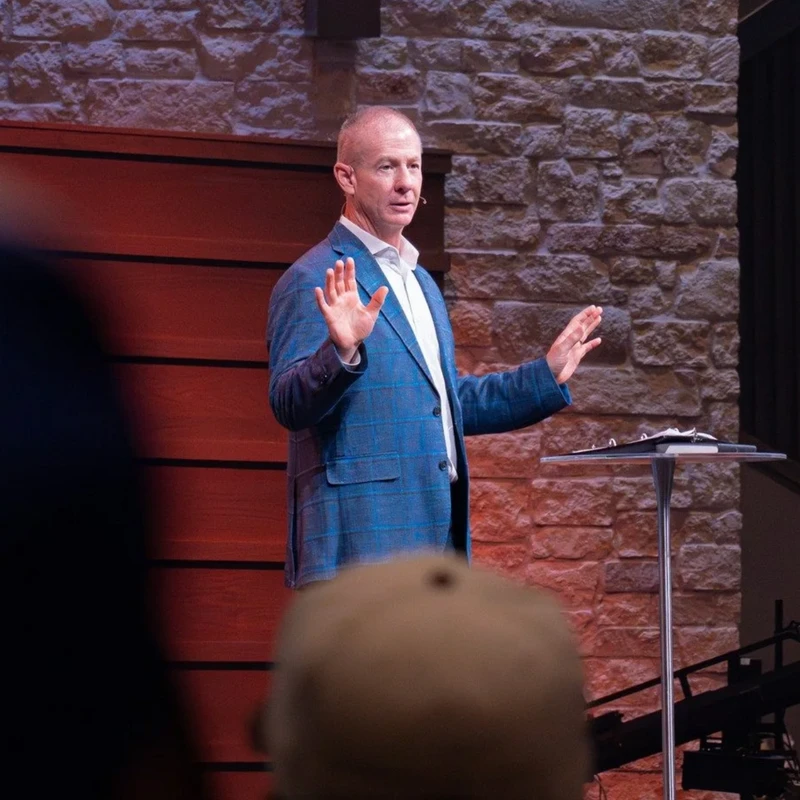 A man with short hair in a blue plaid blazer speaks on stage. He stands with his hands raised in front of a wood and stone wall, with an audience in front.