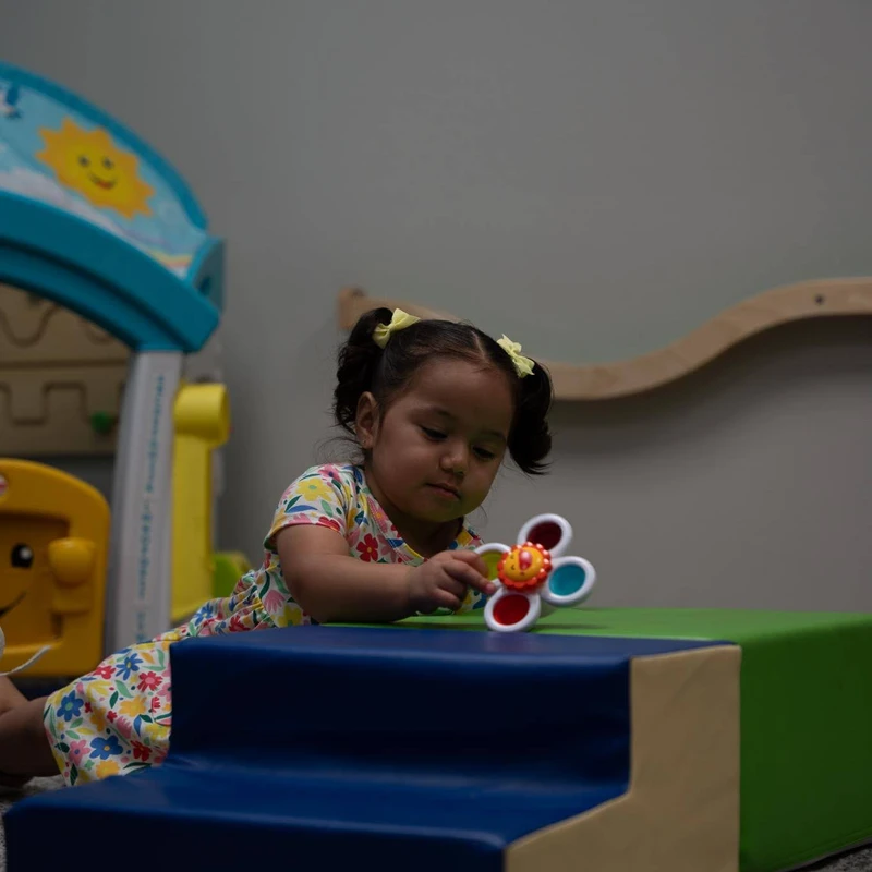 A young girl with pigtails in a floral dress leans on a soft play block, intently focused on a colorful, flower-shaped spinner toy she is holding.