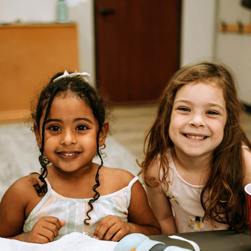 Two diverse young girls sit side-by-side at a table in what appears to be a classroom. They are both looking at the camera and smiling happily.