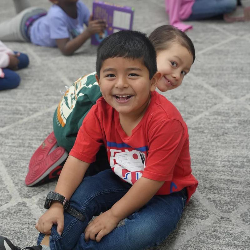 A young boy in a red shirt smiles brightly at the camera while sitting on a classroom floor. Another boy playfully peeks out from behind him.