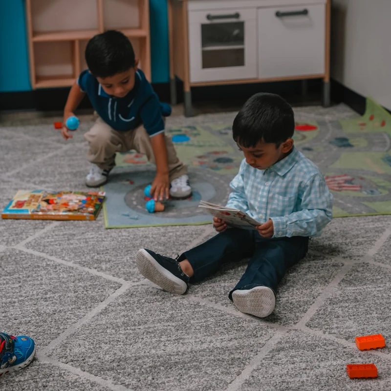 Two young boys on a gray carpet in a playroom. One boy sits reading a book while the other kneels in the background playing with toys near a toy kitchen.