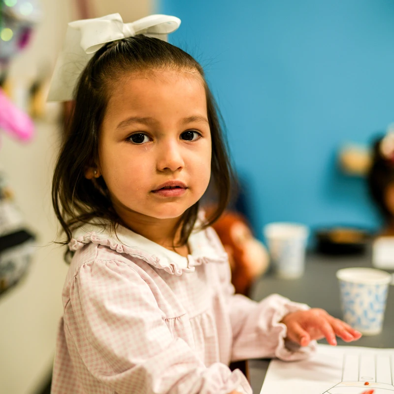 A young girl with a big white bow in her hair sits at a desk in a classroom, looking at the camera while holding an orange crayon over a coloring page.