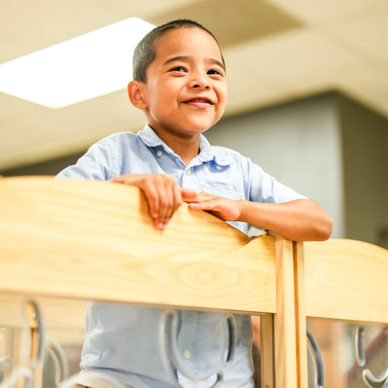 A low-angle shot of a young boy with dark hair smiling warmly. He is wearing a blue shirt and resting his hands on a wooden railing in an indoor setting.