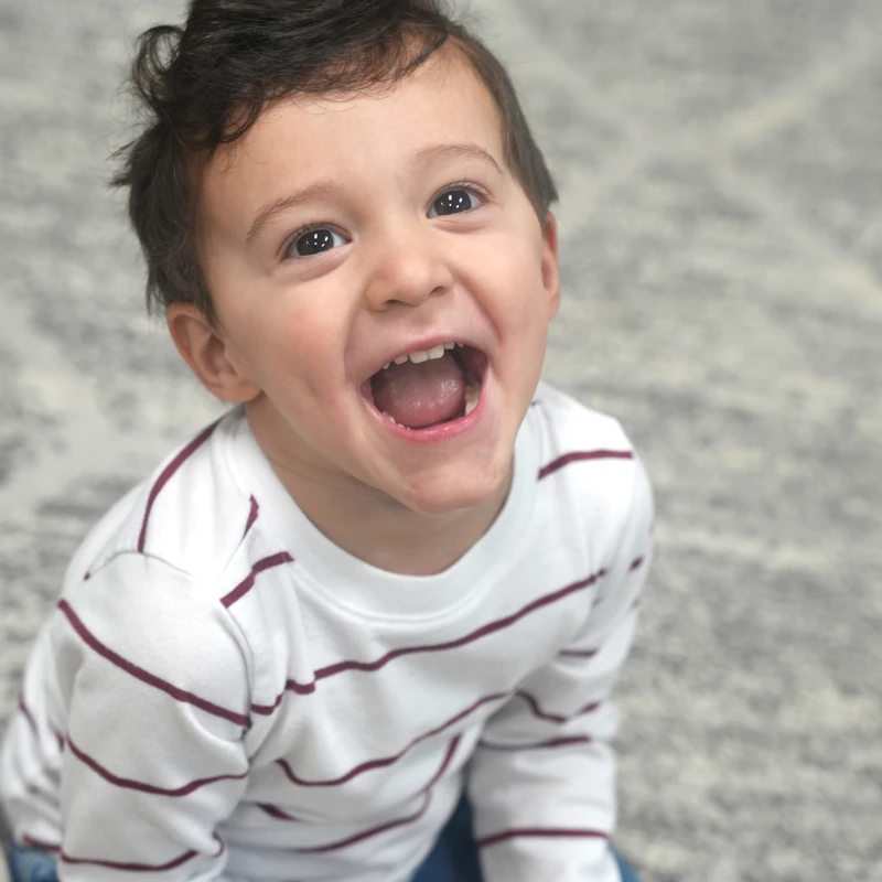 A high-angle portrait of a happy toddler with dark hair and a striped shirt, looking up at the camera with a wide, joyful, open-mouthed smile.