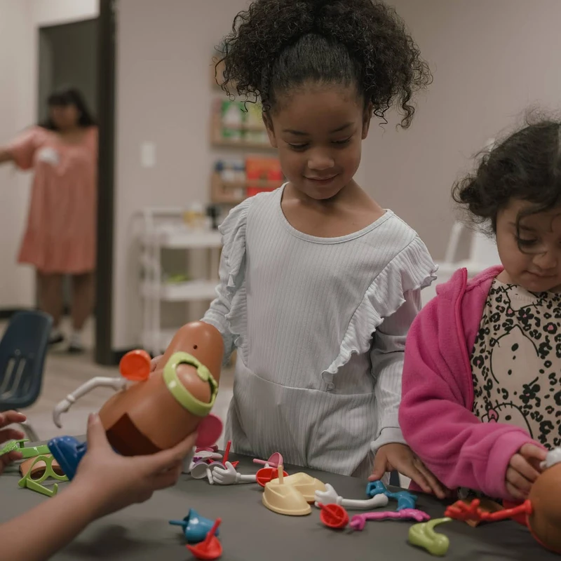 Two young girls stand at a table in a classroom playing with a Mr. Potato Head toy, with colorful plastic pieces scattered around them.