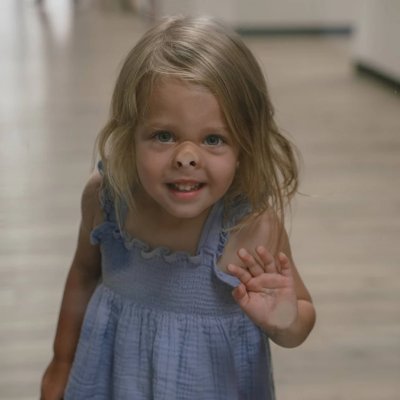 A young girl with blonde hair in a blue dress smiles and waves while pressing her nose against a pane of glass, looking at the camera.
