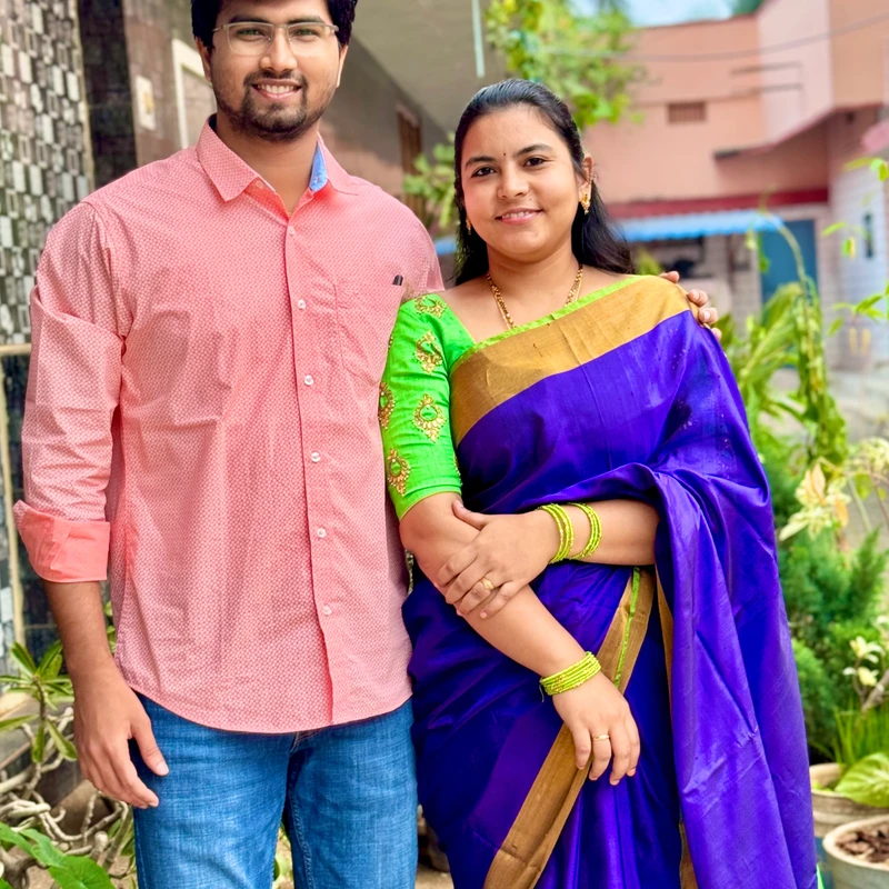 A man in a pink shirt and jeans stands with his arm around a woman in a vibrant purple and green sari. They are both smiling for the camera in a garden.