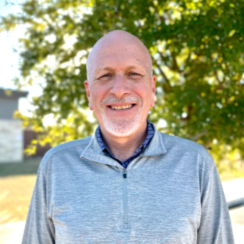 A head-and-shoulders portrait of a smiling bald man with a gray beard, wearing a gray pullover outdoors in front of a leafy green tree.