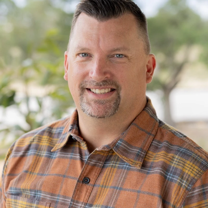 A headshot of a smiling, middle-aged white man with blue eyes and a goatee. He is wearing an orange, yellow, and blue plaid shirt and is outdoors.