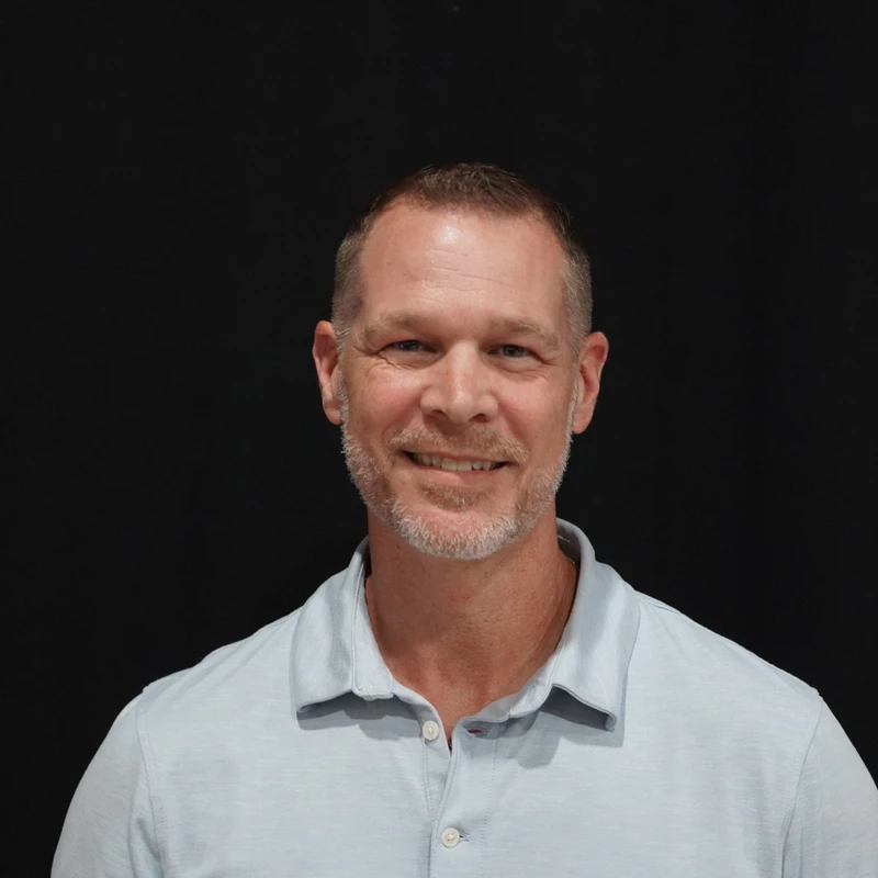 A head-and-shoulders portrait of a smiling middle-aged man with short hair and a goatee, wearing a light blue polo shirt against a black background.