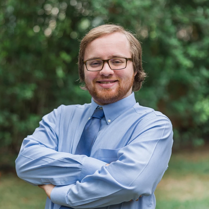 A friendly-looking man with reddish-blond hair, a beard, and glasses smiles with his arms crossed. He wears a light blue shirt and a tie outdoors.
