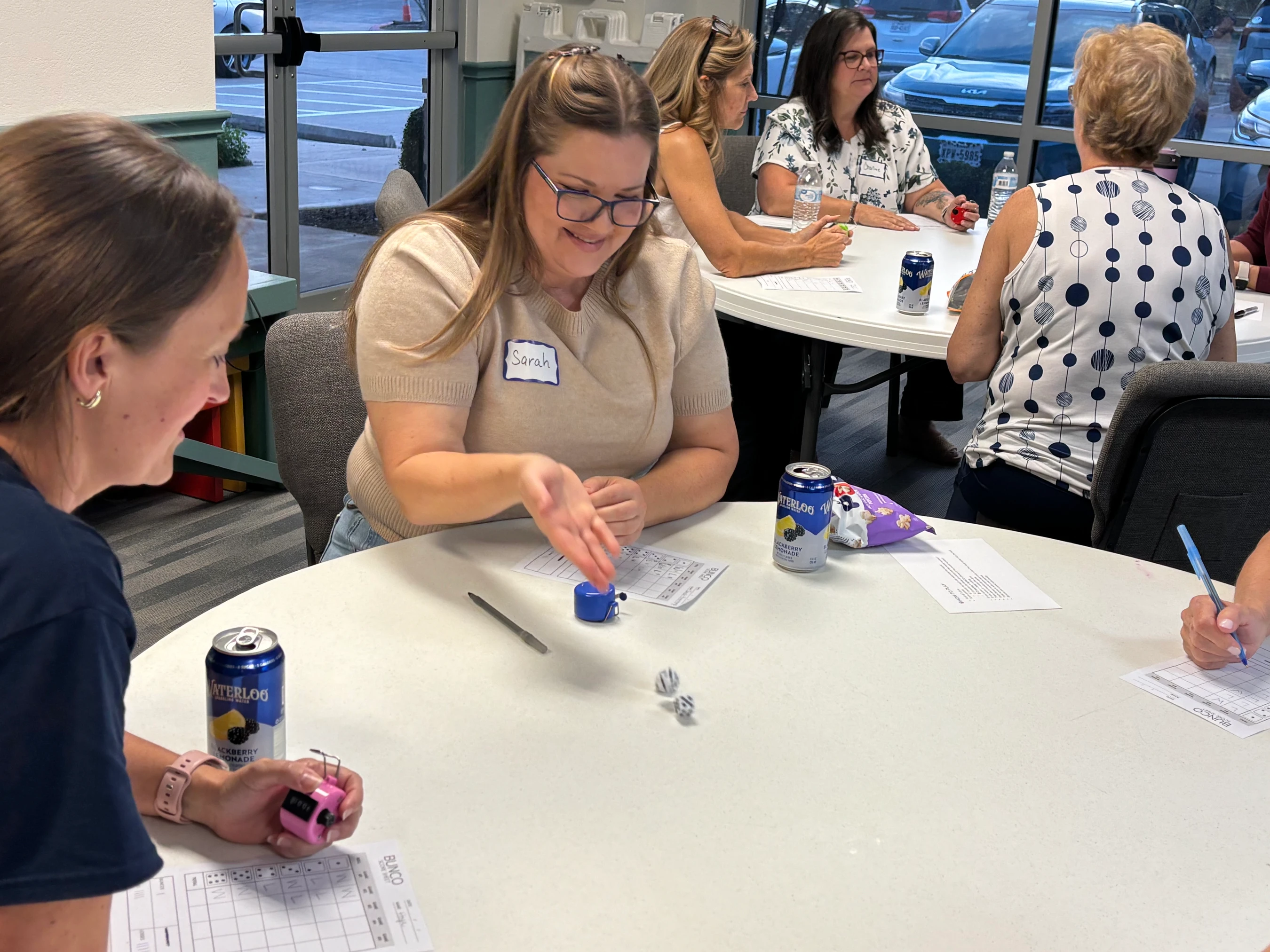 A group of women sit at white tables playing the dice game Bunco. A woman named Sarah smiles as she drops dice onto the table during her turn.