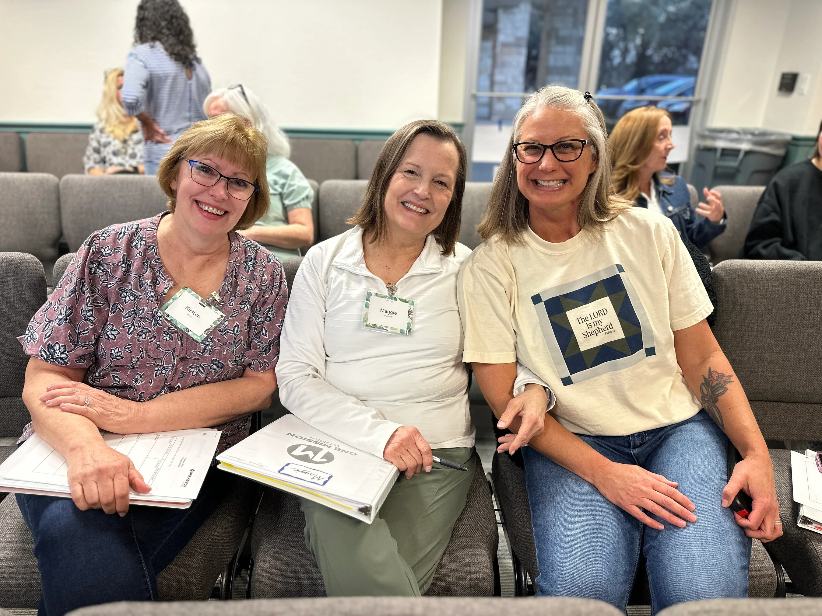 Three women sitting next to each other in gray chairs at a conference or event. They are all smiling at the camera and holding binders in their laps.