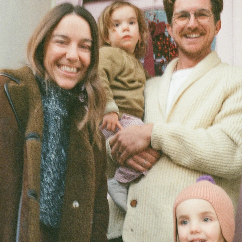 A grainy, film-style photo of a smiling family of four standing outside their front door. The father holds a toddler, and another young girl is in front.