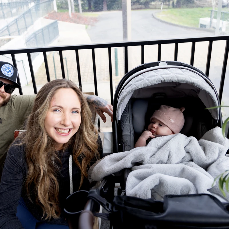 A man, a woman, and their baby in a car seat smile for the camera while sitting on a balcony. The man wears a cap and sunglasses.