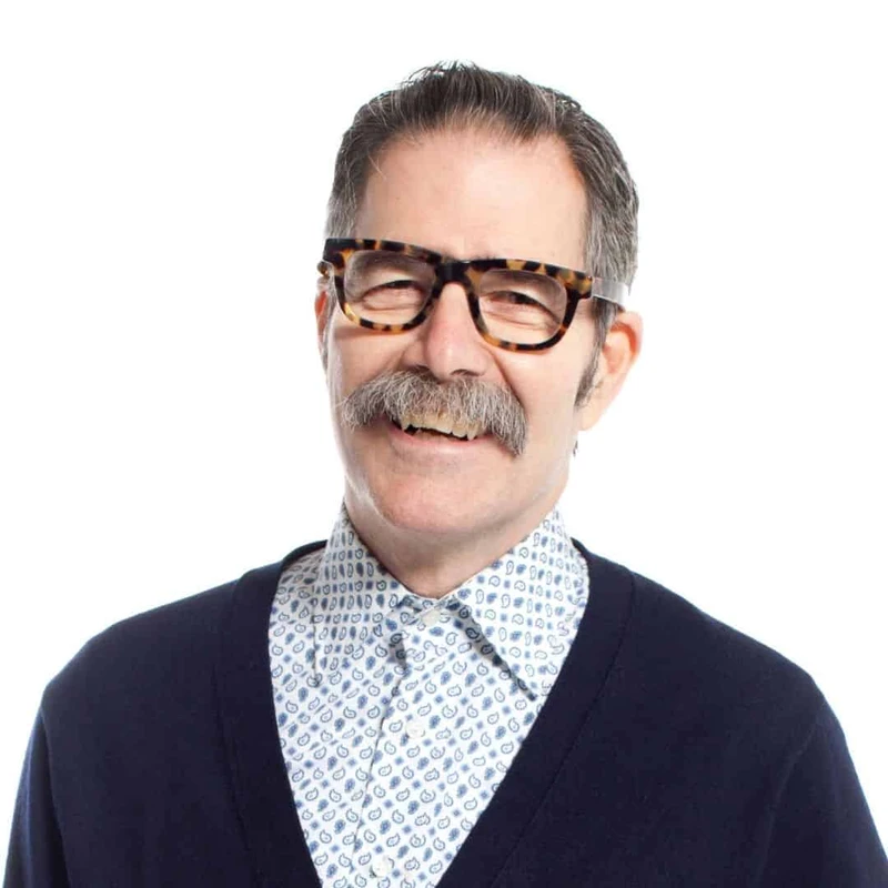 A headshot of a smiling man with a gray mustache and thick tortoiseshell glasses, wearing a patterned shirt and a dark blue cardigan against a white background.