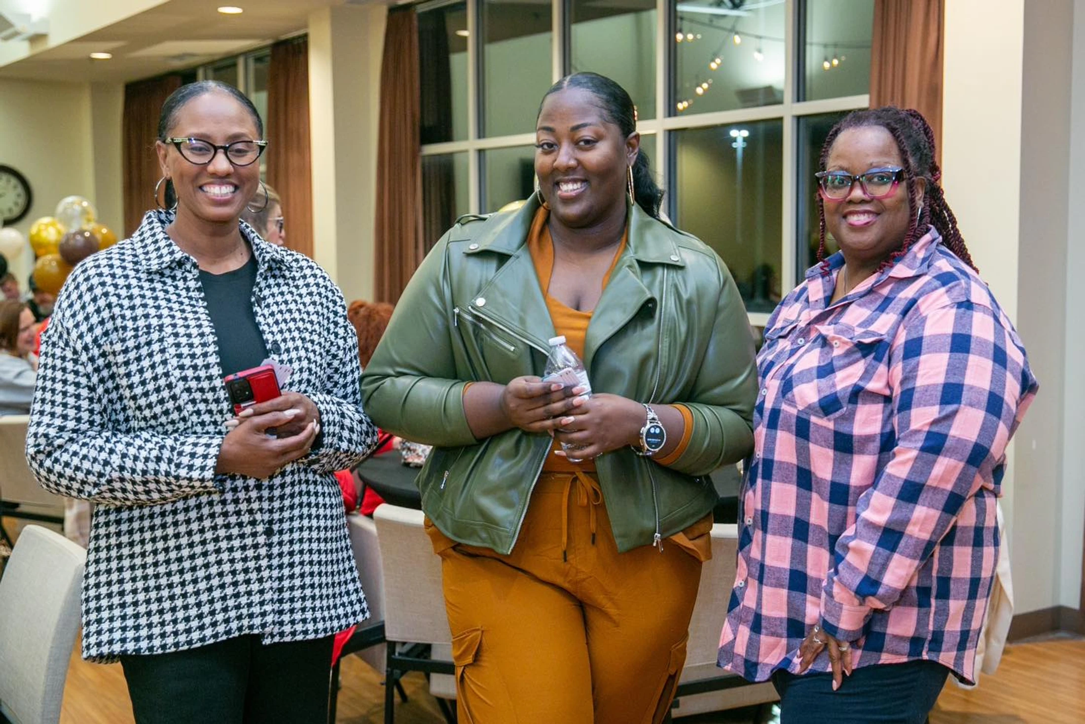 Three smiling Black women stand together for a photo at an indoor gathering. They are wearing a houndstooth jacket, a green leather jacket, and a plaid shirt.