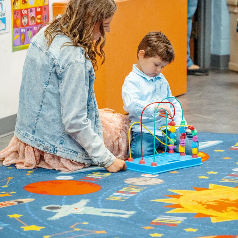 A woman and a young boy sit on a colorful space-themed rug. The boy is focused on playing with a bead maze toy while the woman watches him.