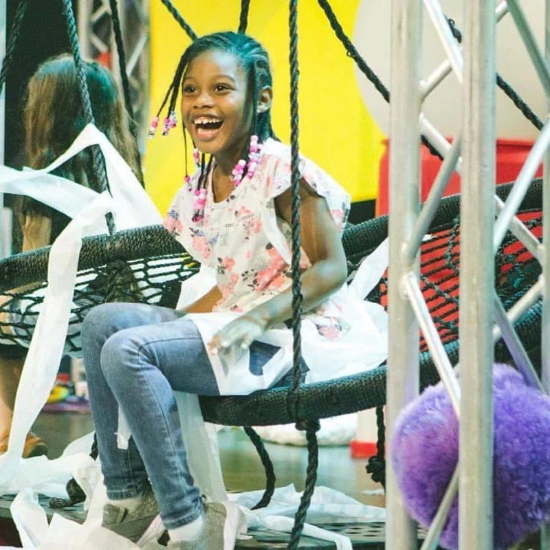 A young girl with braided hair and pink beads smiles happily while sitting on a large, round, black woven swing covered in white streamers indoors.