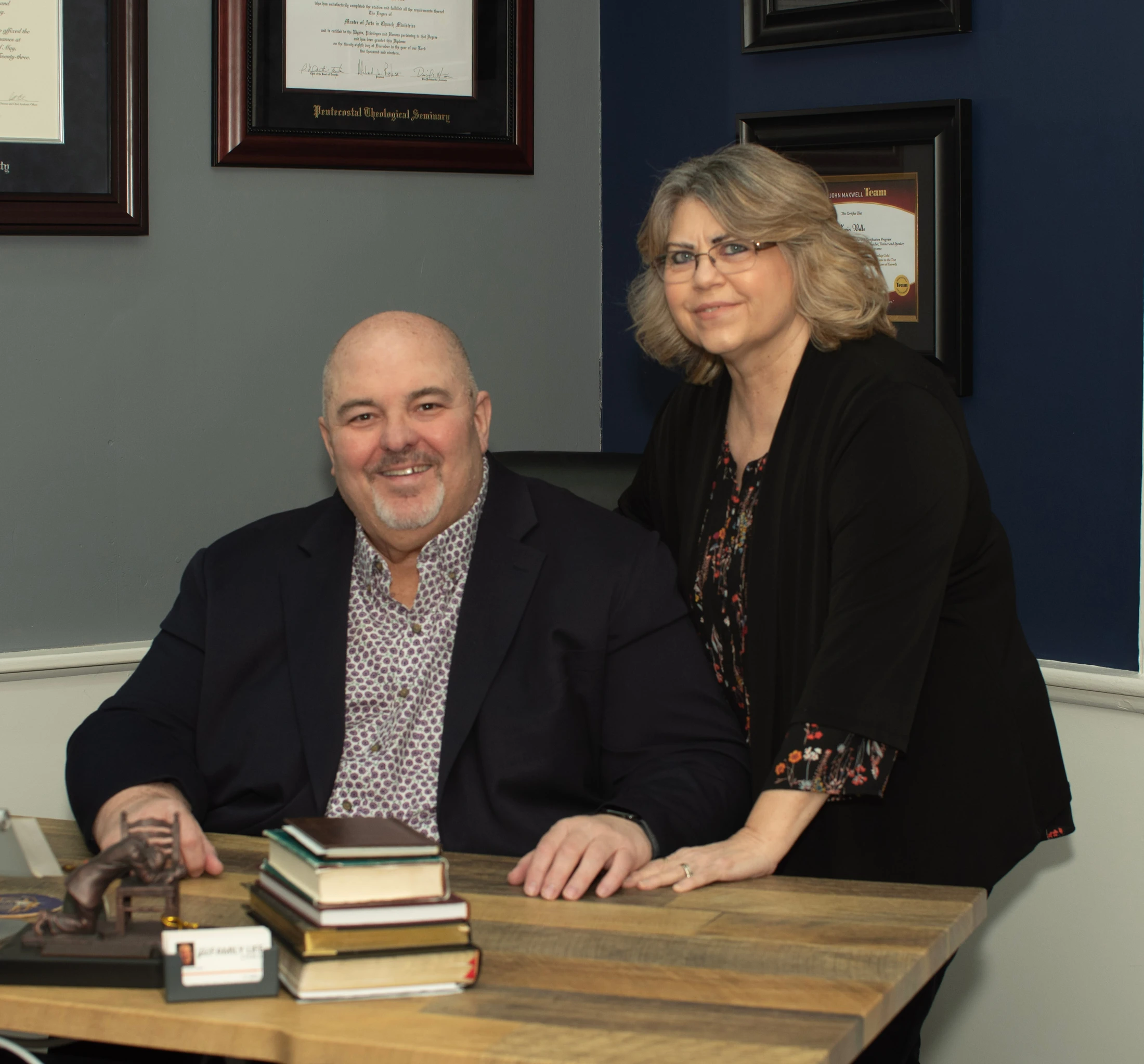 A smiling man sits at a wooden desk in an office, while a woman in glasses stands behind him with her hand on the desk. Both look at the camera.