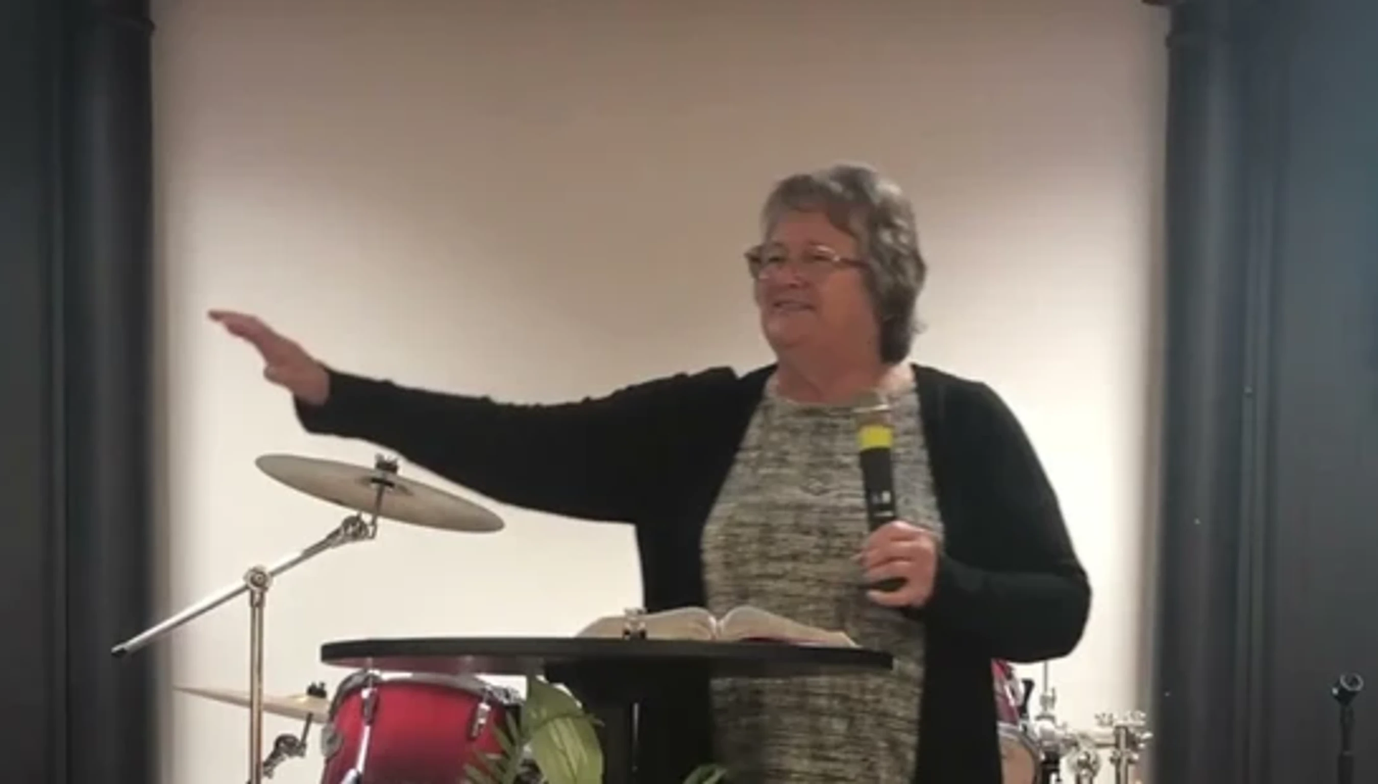 A woman with short hair and glasses wearing a black cardigan speaks into a microphone while standing at a podium on a stage with a drum set behind her.