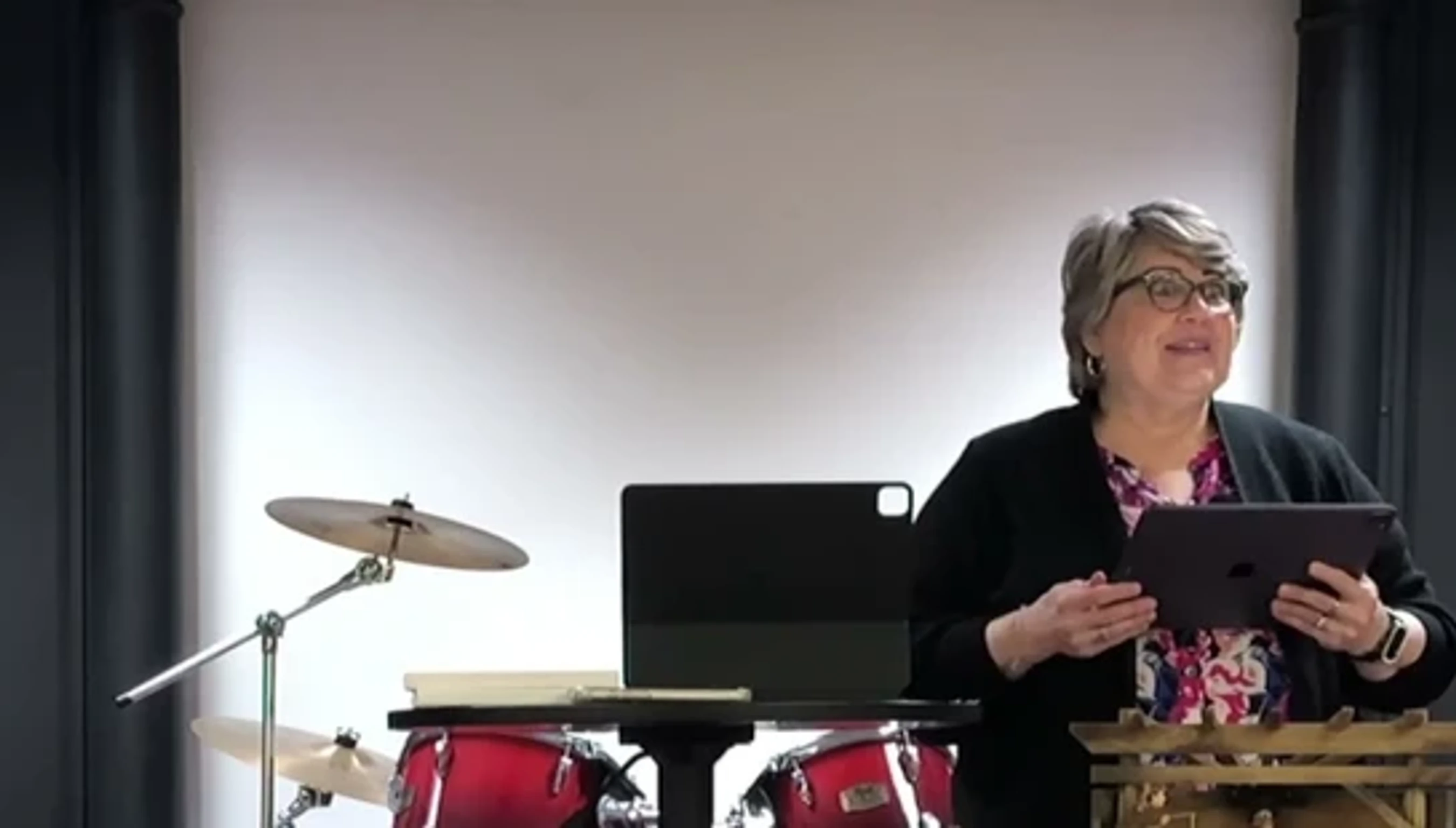 A woman with short gray hair and glasses holds a tablet, speaking on a stage with a red drum kit behind her against a white wall.