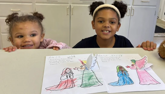 Two young girls smile as they peek over a table, showing off their colored pop-up drawings of an angel visiting Mary from a Bible story.