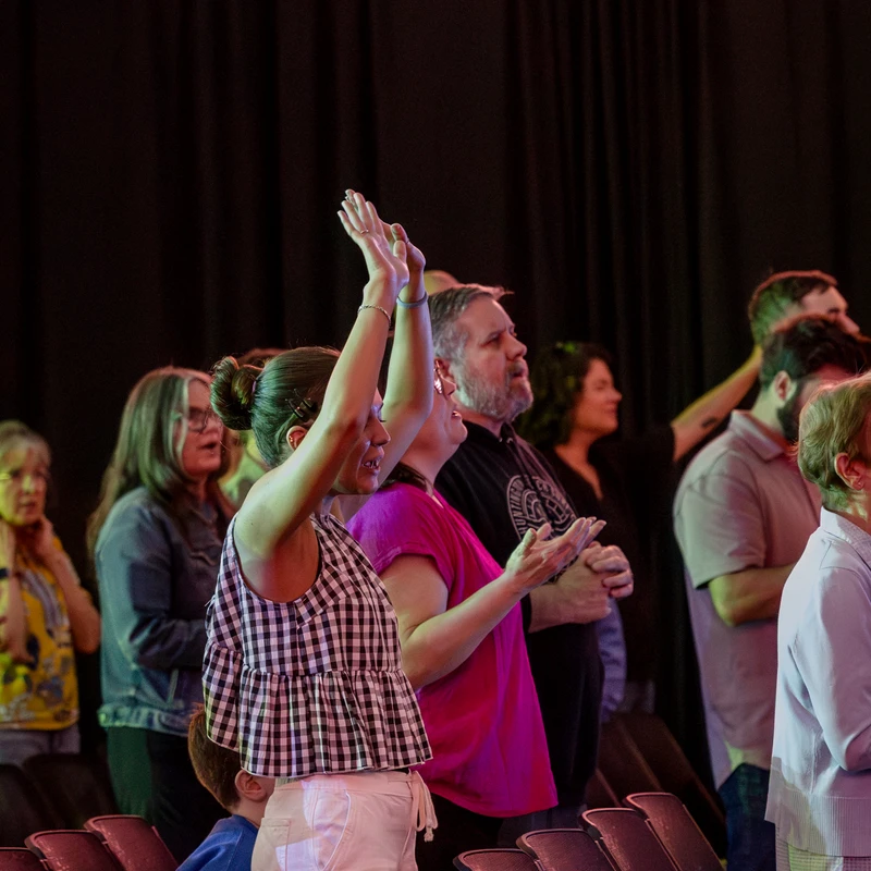 A diverse group of people stand in rows of chairs during a service. A woman in a checkered top is in focus, raising her hands in an act of praise.