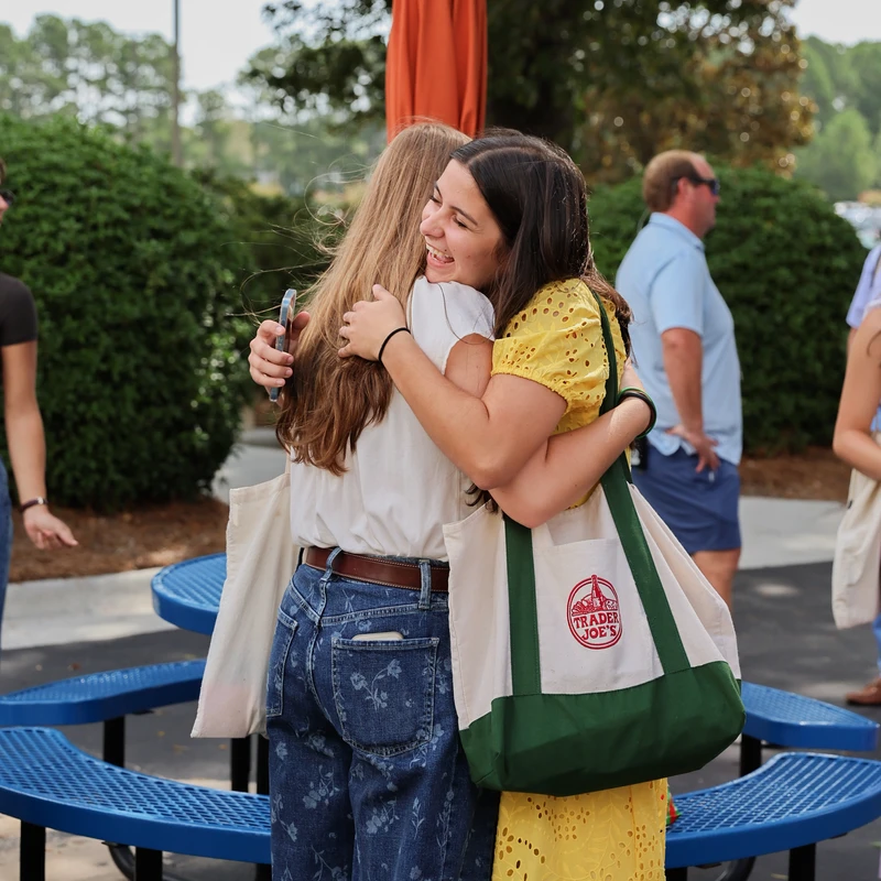 Two young women share a happy hug outdoors near a blue picnic table. One woman in a yellow dress and carrying a Trader Joe's bag is smiling broadly.