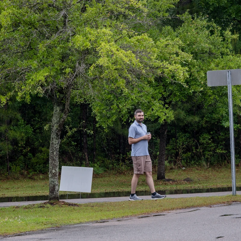 A man in a blue polo shirt and shorts walks on a sidewalk through a park. He holds a cup, and the background is filled with lush green trees and grass.