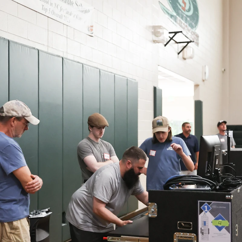 A group of men set up audio-visual equipment from a large black road case inside a school gym, with a Holly Shelter Middle School banner on the wall.