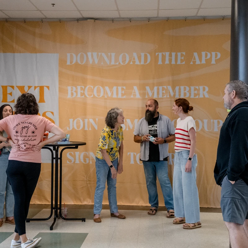 A group of adults talking and laughing in a hallway in front of a large yellow banner that reads "Next Steps" and "Become a Member."