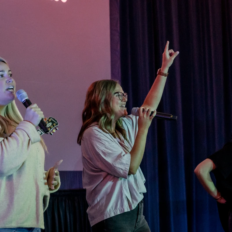 Three women sing into microphones on a stage with purple lighting. The woman in the center raises her hand in the air while singing with enthusiasm.