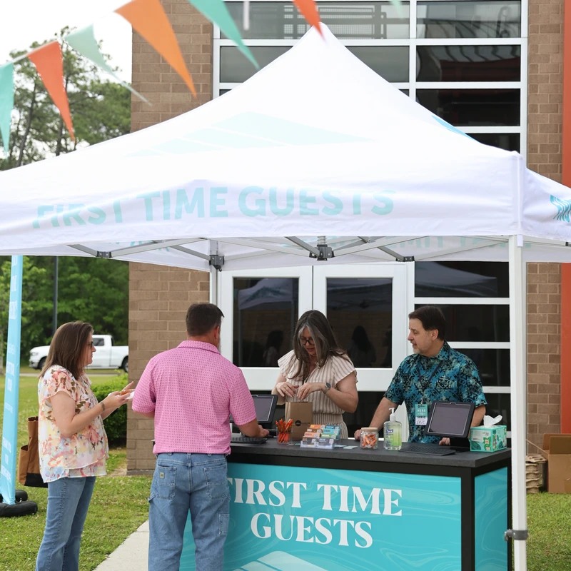 Volunteers welcome a couple at a 'First Time Guests' booth under a white tent. Colorful pennant flags hang overhead outside a brick building.