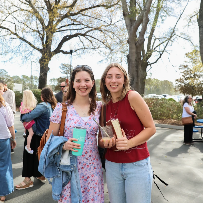 Two young women stand side-by-side and smile at the camera. They are outdoors in a parking lot on a sunny day, with trees and other people in the background.