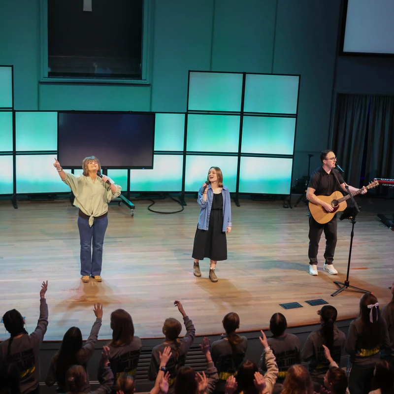 A worship band performs on stage for an audience of young people, who stand with their hands raised in praise. A large cross is visible on the wall.