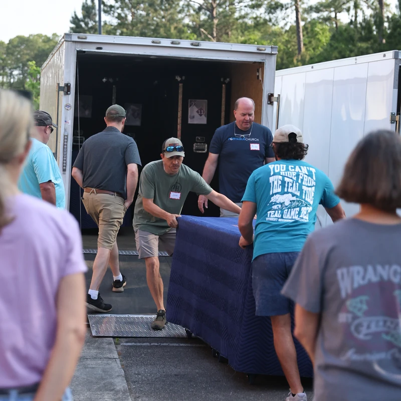 A group of people work together to move a large object covered in a blue blanket down the ramp of a white truck trailer in a parking lot.